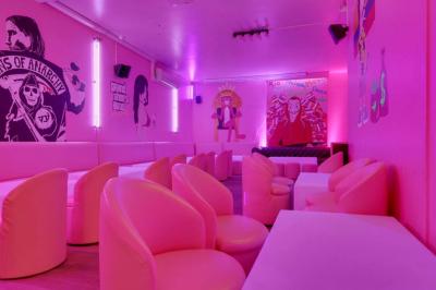 Salle de bar aux murs roses avec banquettes blanches, tabourets assortis et néons décoratifs.