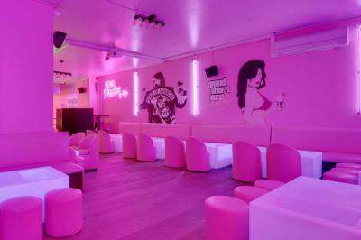 Salle de bar aux murs roses avec banquettes blanches, tabourets assortis et néons décoratifs.
