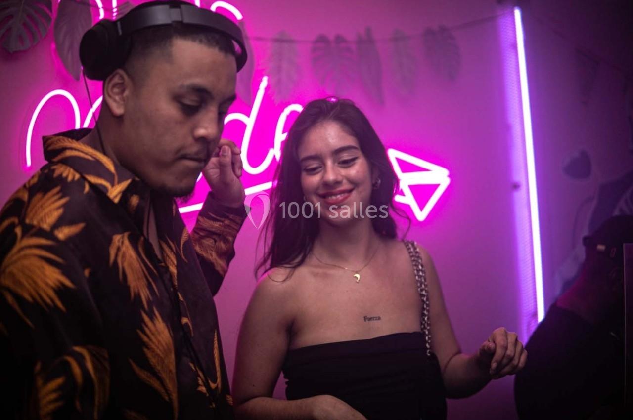 Un DJ mixe de la musique tandis qu'une femme sourit dans une ambiance tamisée avec des néons roses et violets.