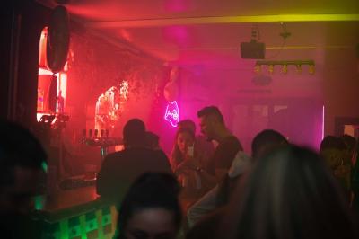Salle de bar aux murs roses avec banquettes blanches, tabourets assortis et néons décoratifs.
