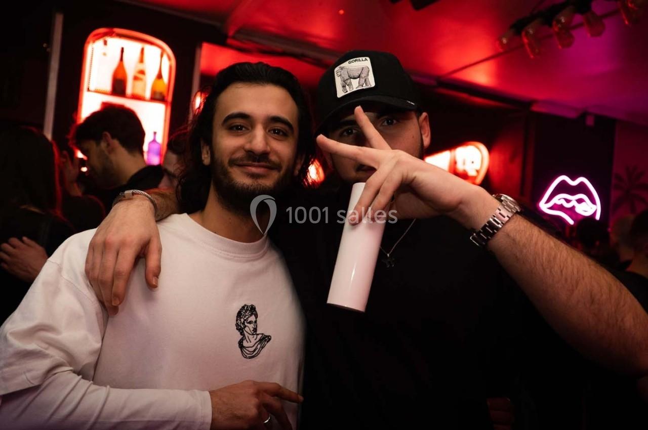 Deux hommes posent dans un bar éclairé par des néons, l'un tenant un gobelet et faisant un signe de la main.