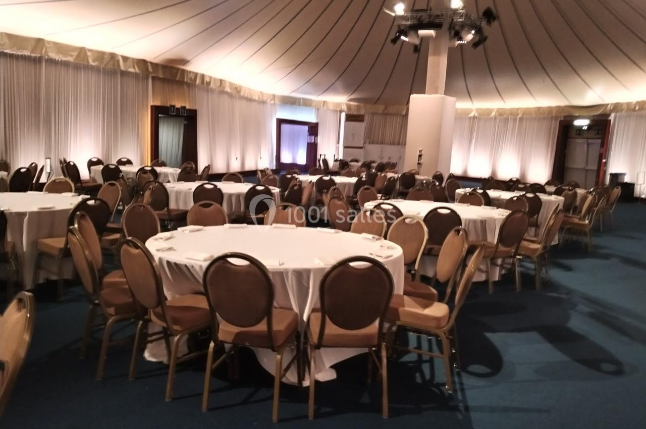 Salle de réception avec des tables rondes dressées de nappes blanches et entourées de chaises marron.