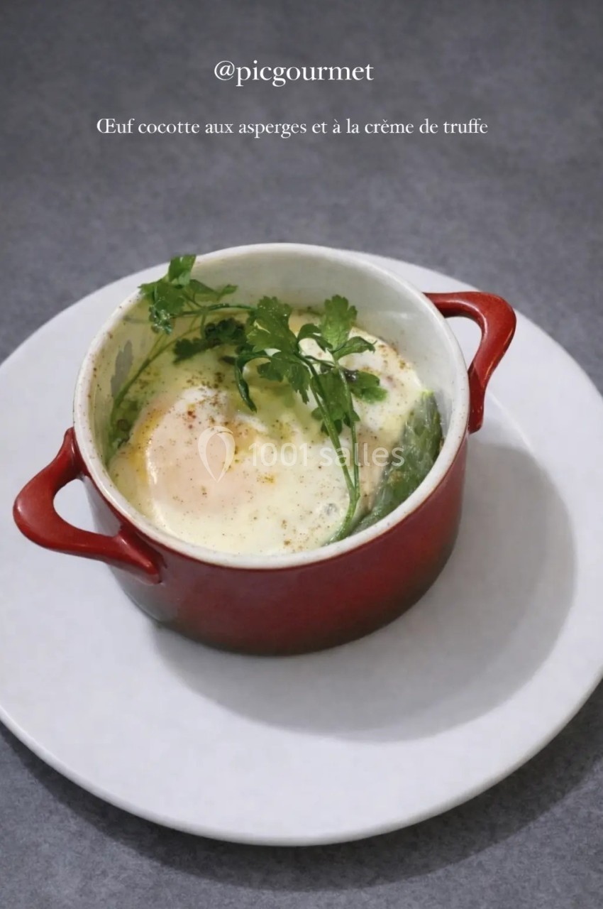 Œuf cocotte dans une petite cocotte rouge, accompagné d'asperges et garni de crème de truffe et de persil.