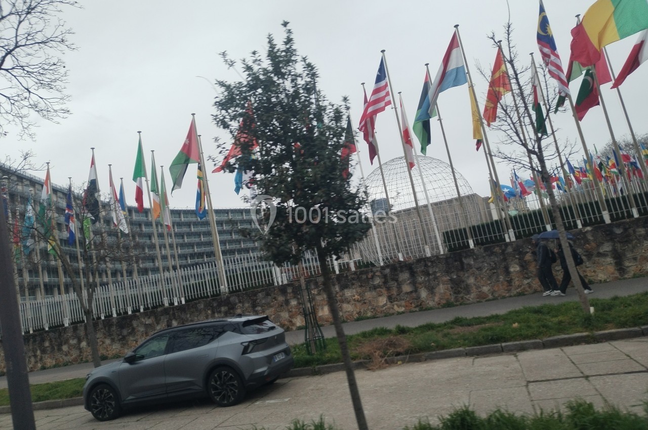 De nombreuses drapeaux internationaux flottent devant un bâtiment entouré de grilles, avec une voiture stationnée au premier…
