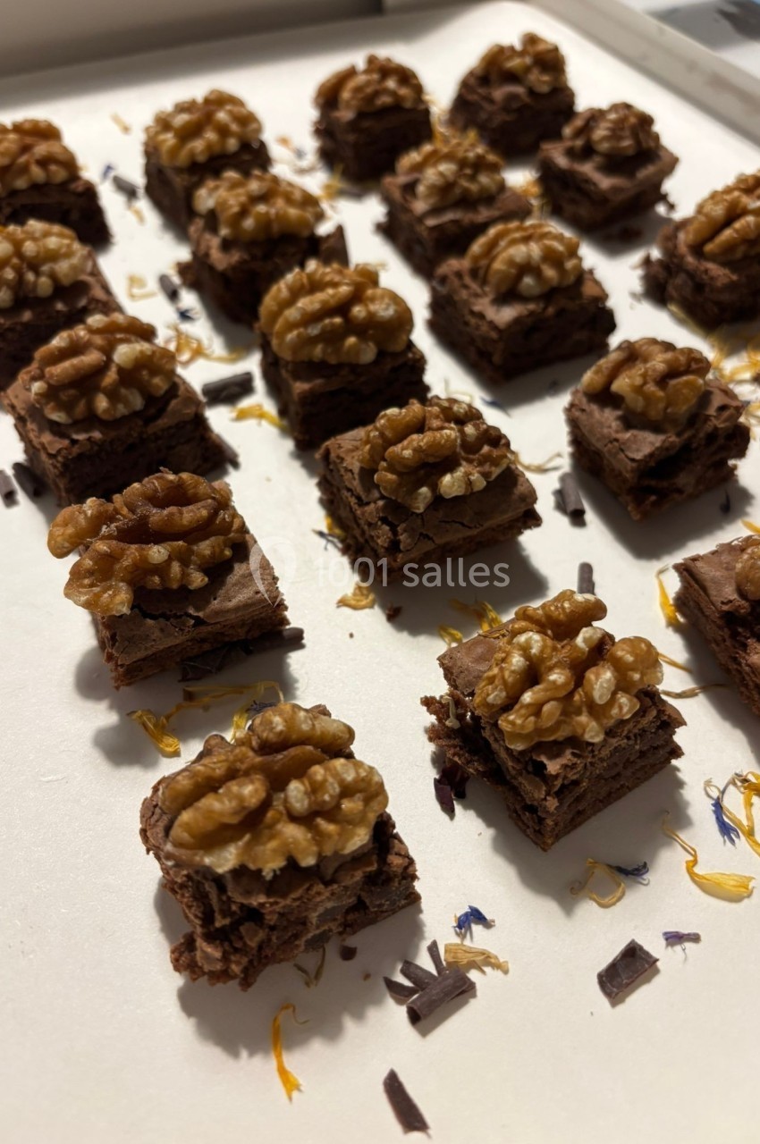 Carrés de gâteau au chocolat garnis de noix, disposés sur un plateau avec des éclats de chocolat et des pétales décoratifs.