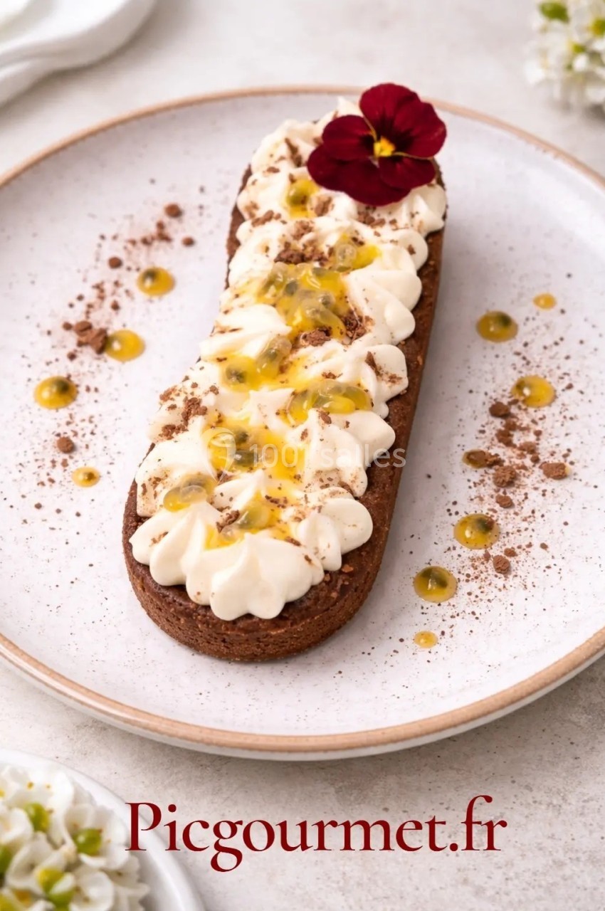 Tartelette au chocolat garnie de crème, fruit de la passion et décorée d'une fleur comestible sur une assiette blanche.