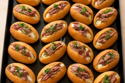 Plateau de petits pains garnis de fromage fondu, viande et persil, disposés en rangées sur une plaque noire.