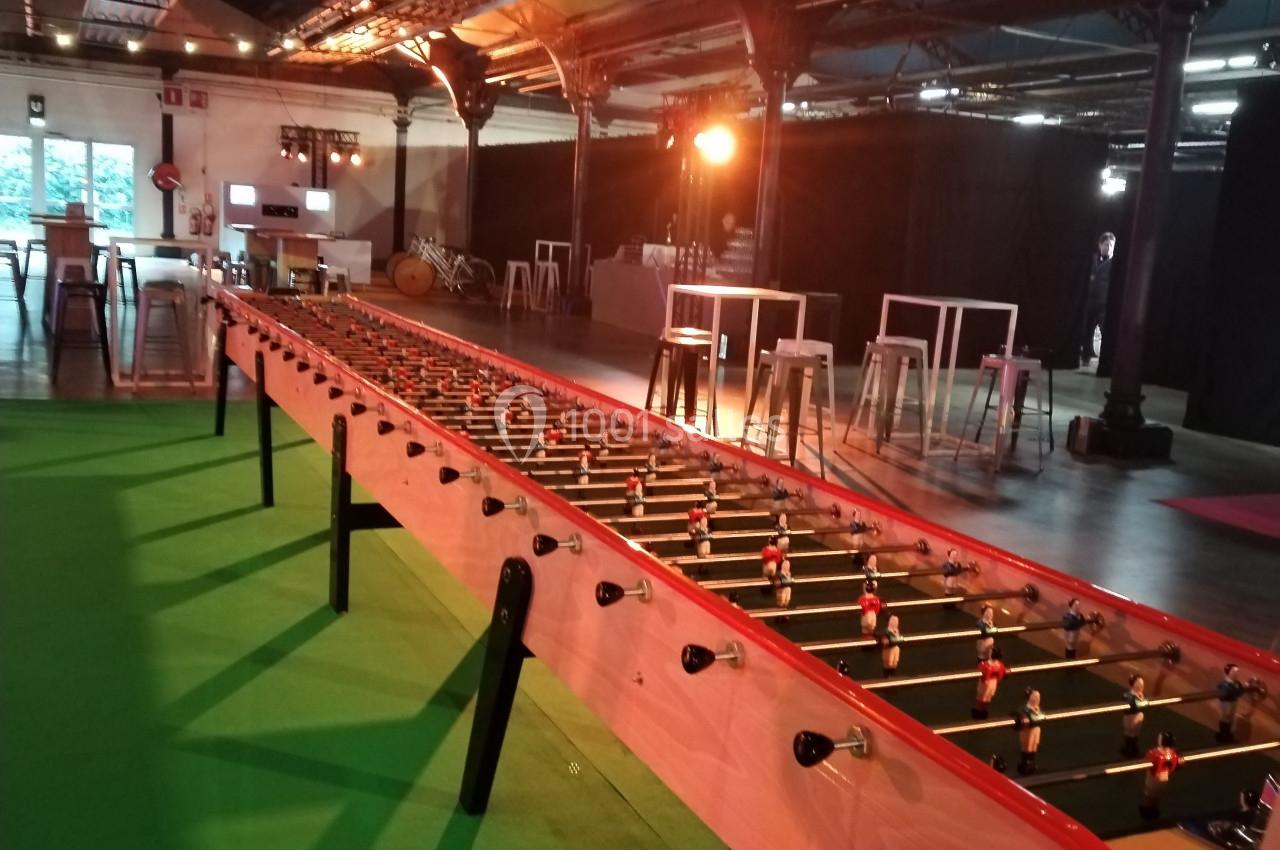 Baby-foot géant installé dans un espace intérieur avec éclairage tamisé et mobilier en arrière-plan.