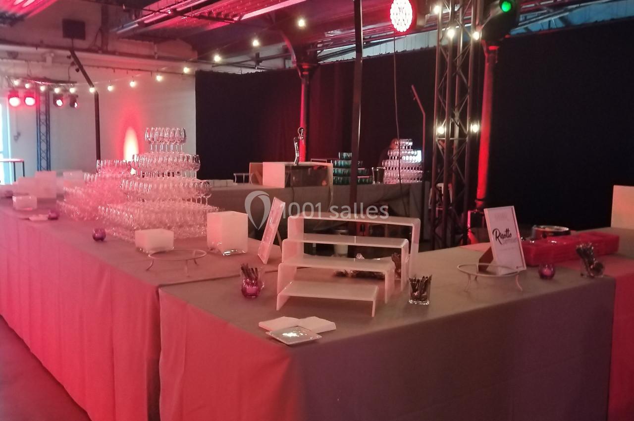 Buffet avec tables dressées, verres empilés, présentoirs blancs et éclairage rouge dans une salle événementielle.