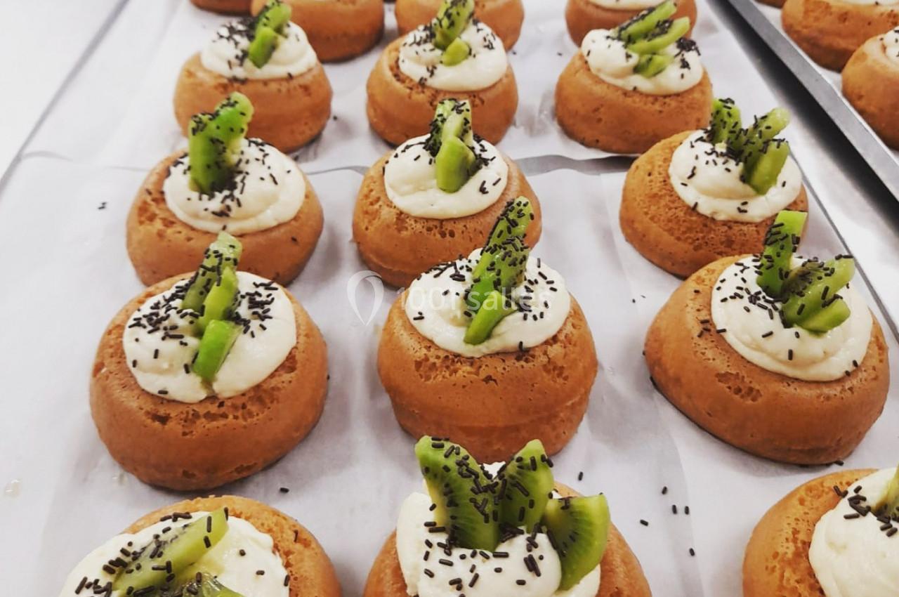 Petits gâteaux individuels garnis de crème et de morceaux de kiwi, décorés de graines noires sur du papier cuisson.