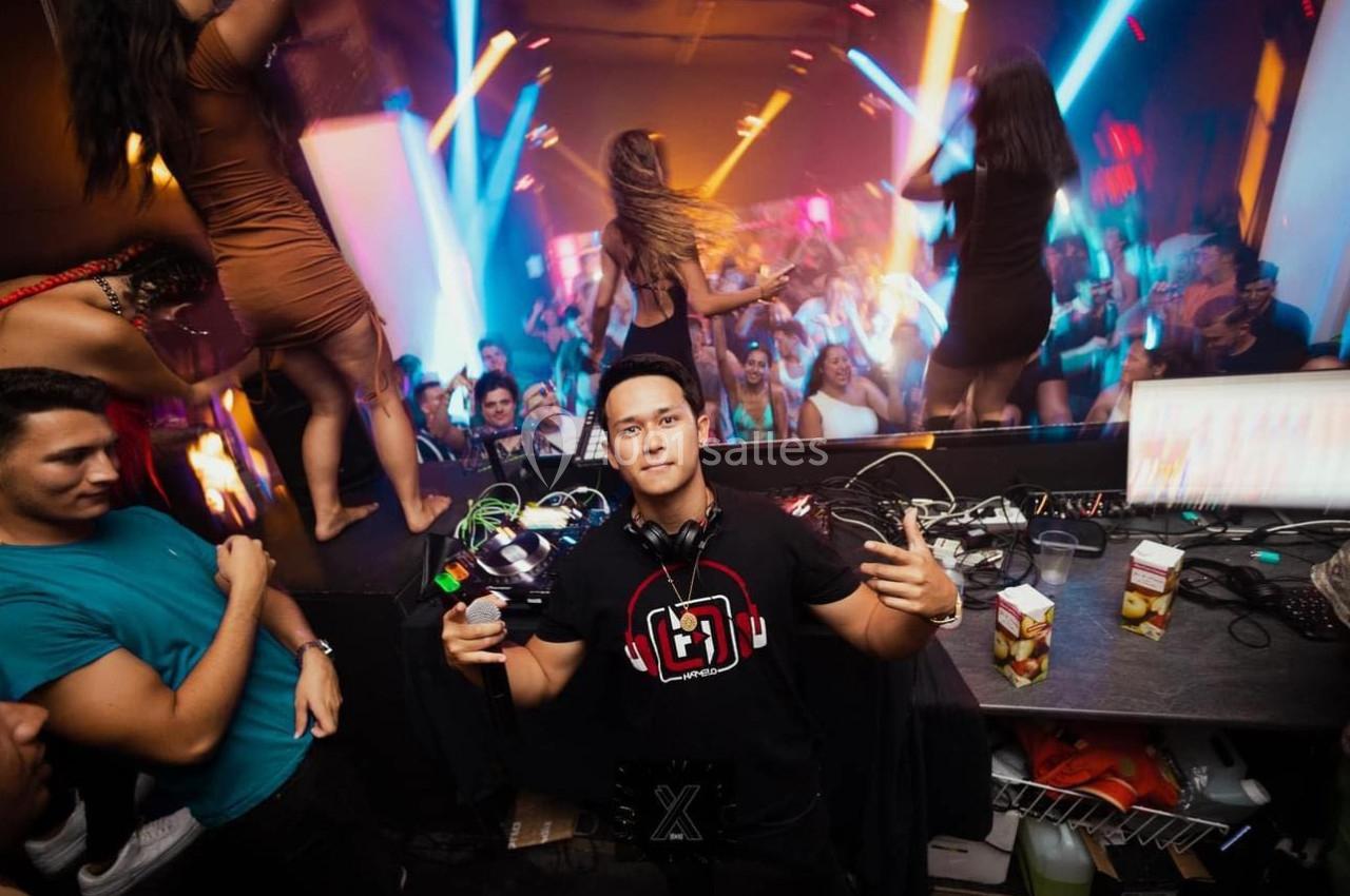 Un DJ pose devant son équipement dans une boîte de nuit animée avec des lumières colorées et une foule dansante.