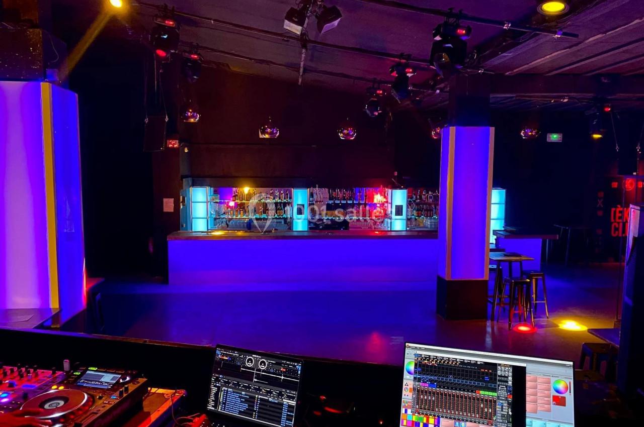 Salle de club avec piste de danse vide, bar éclairé par des lumières colorées et équipement de DJ au premier plan.