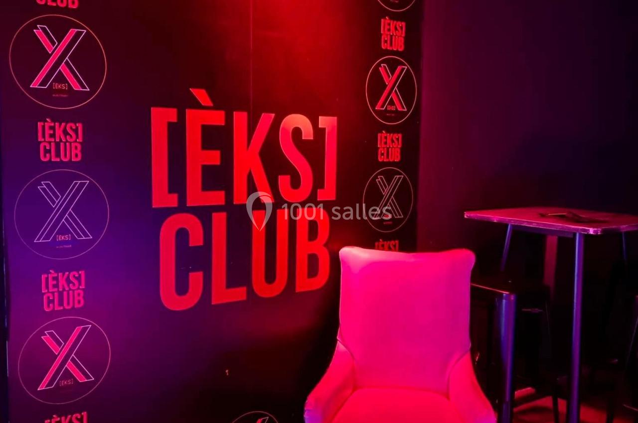 Fauteuil rose devant un mur noir avec le logo ’[ÈKS] CLUB’ illuminé par une lumière rouge.