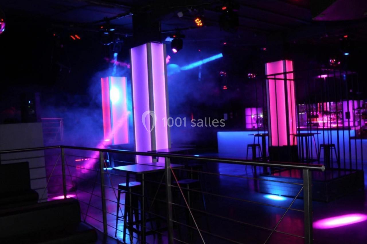 Salle de discothèque avec éclairage coloré, colonnes lumineuses, bar en arrière-plan et ambiance tamisée.