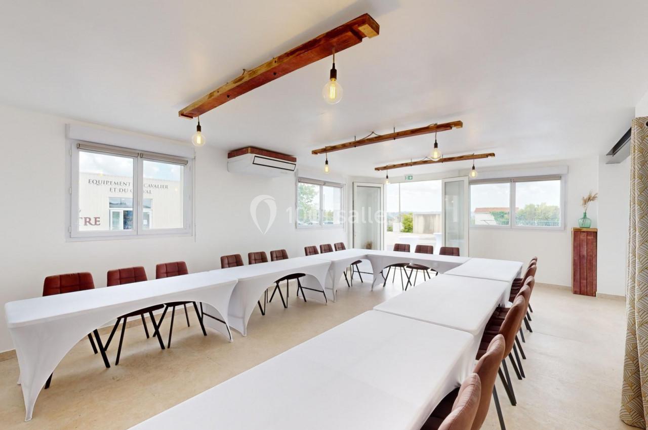 Salle de réunion lumineuse avec tables en U, chaises marron, grandes fenêtres et éclairage suspendu.