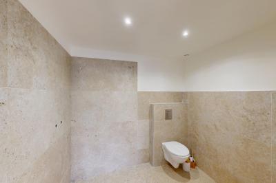 Salle de bain avec murs en carrelage beige, lavabo mural avec accessoires et toilettes séparées par une cloison.