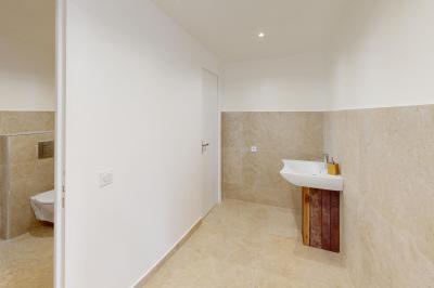 Salle de bain avec murs en carrelage beige, lavabo mural avec accessoires et toilettes séparées par une cloison.