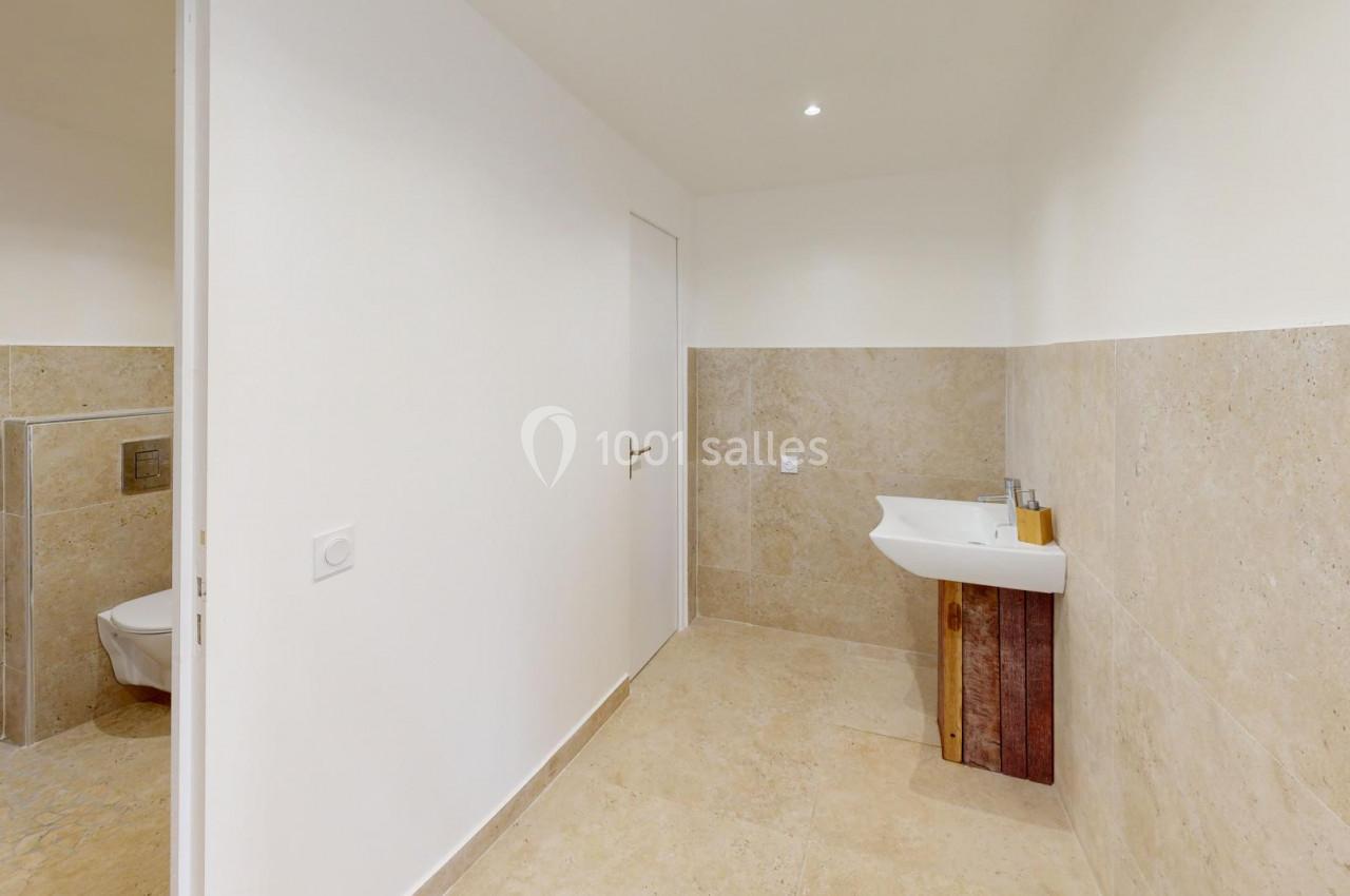 Salle de bain avec murs en carrelage beige, lavabo mural avec accessoires et toilettes séparées par une cloison.