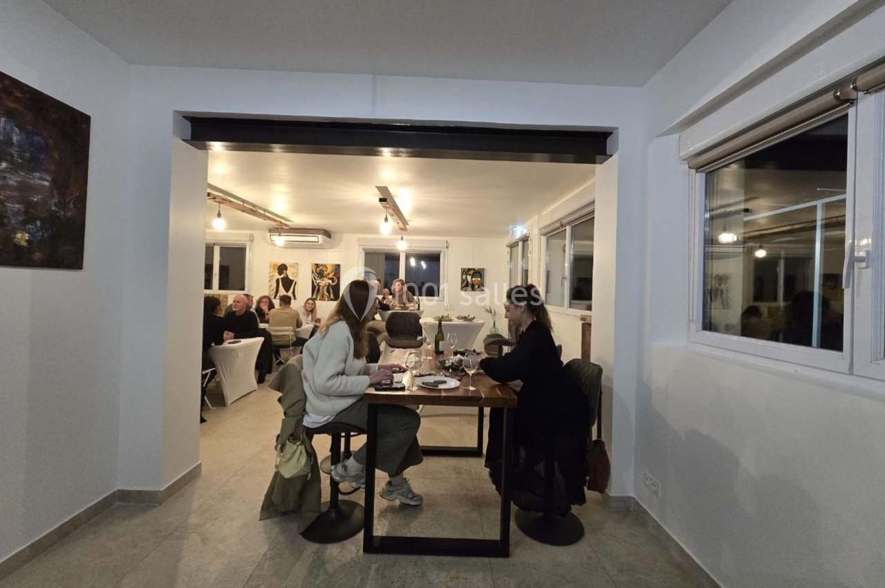 Deux personnes dînent à une table dans une salle lumineuse où d'autres convives sont rassemblés au fond.