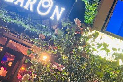 Enseigne lumineuse ’Baron’ entourée de verdure, avec des roses au premier plan et une façade éclairée en arrière-plan.