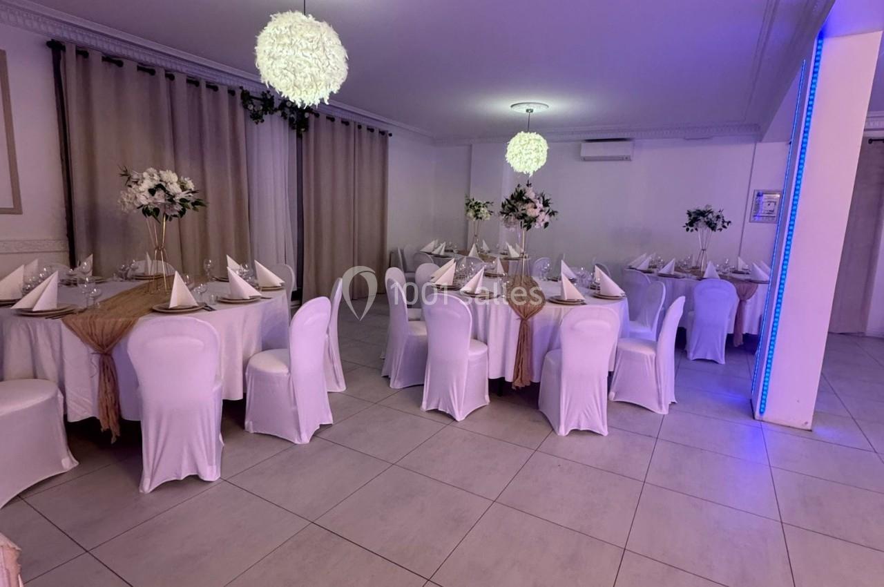 Salle de réception décorée avec des tables nappées, chaises blanches et centres de table floraux sous des luminaires…