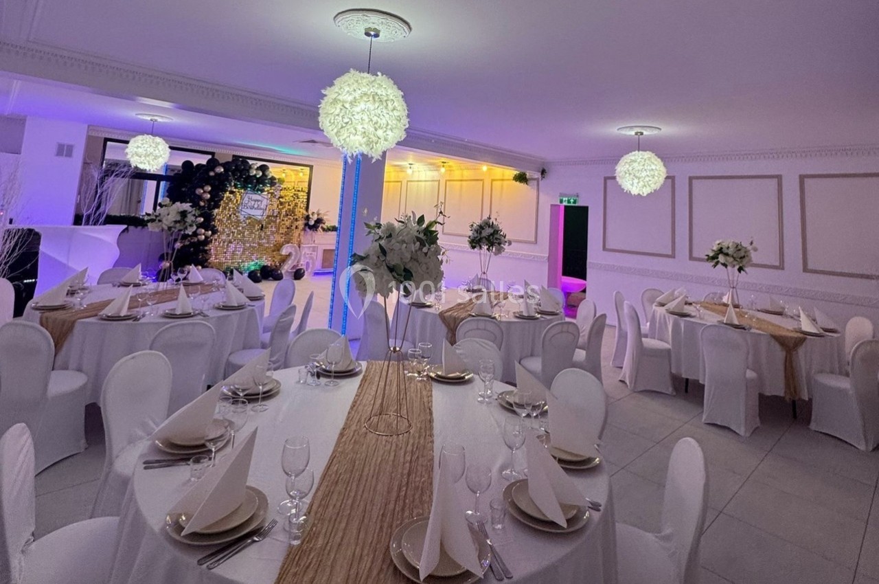 Salle de réception élégamment décorée avec des tables rondes dressées, nappes blanches et éclairage chaleureux.