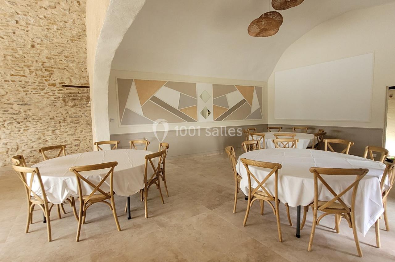 Salle lumineuse avec murs en pierre, tables rondes couvertes de nappes blanches et chaises en bois clair.