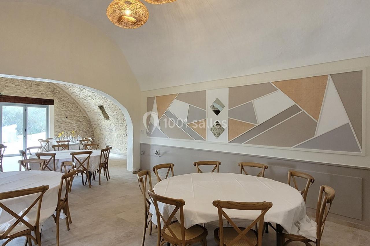 Salle lumineuse avec tables rondes dressées, chaises en bois, murs en pierre et décoration murale géométrique.