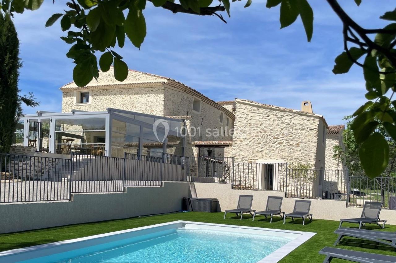 Maison en pierre avec terrasse vitrée, piscine et transats, entourée de végétation sous un ciel bleu.