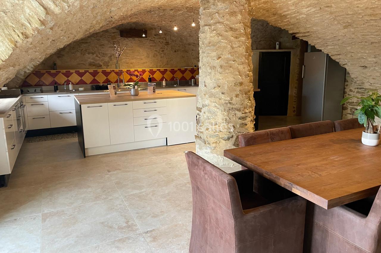Cuisine voûtée en pierre avec meubles modernes, table en bois, chaises marron et crédence colorée.