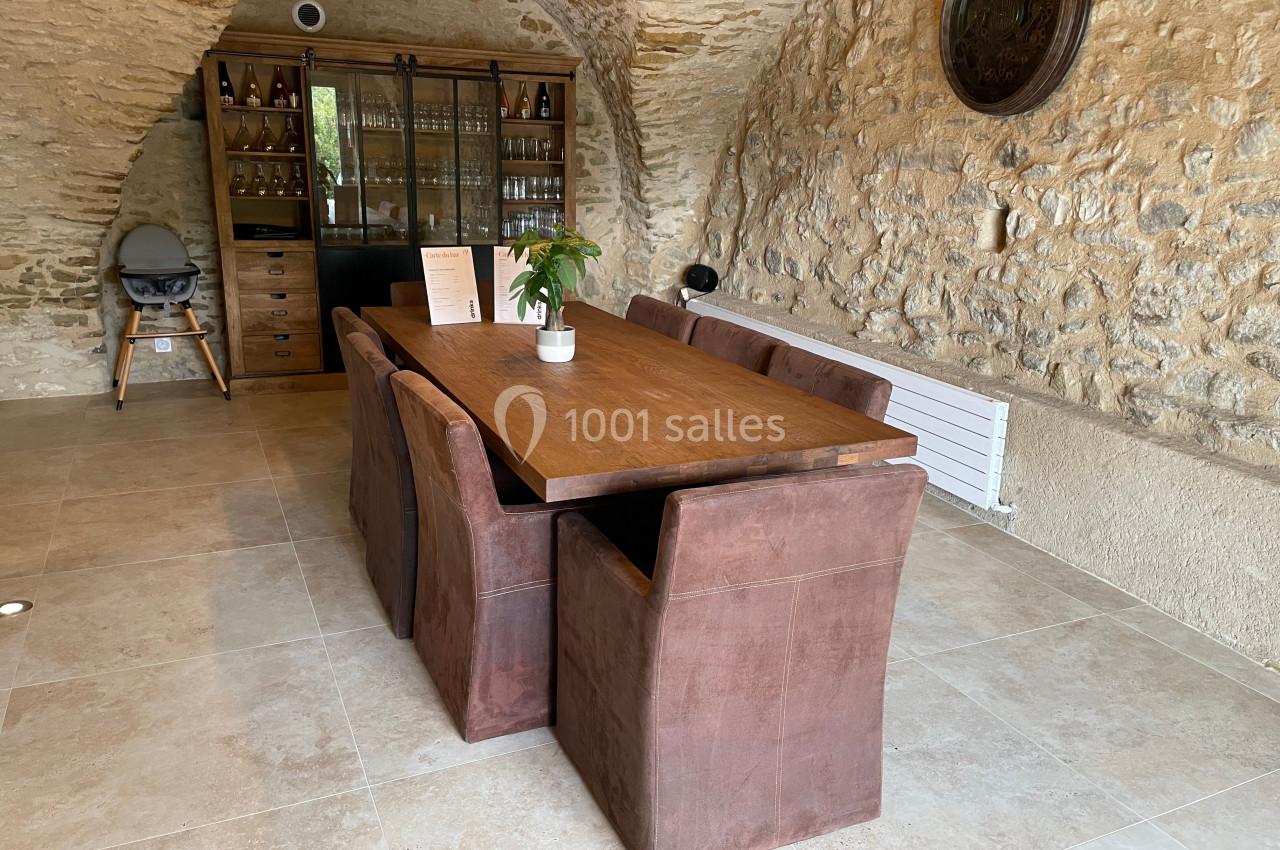 Table en bois entourée de chaises marron dans une pièce en pierre avec un buffet vitré en arrière-plan.