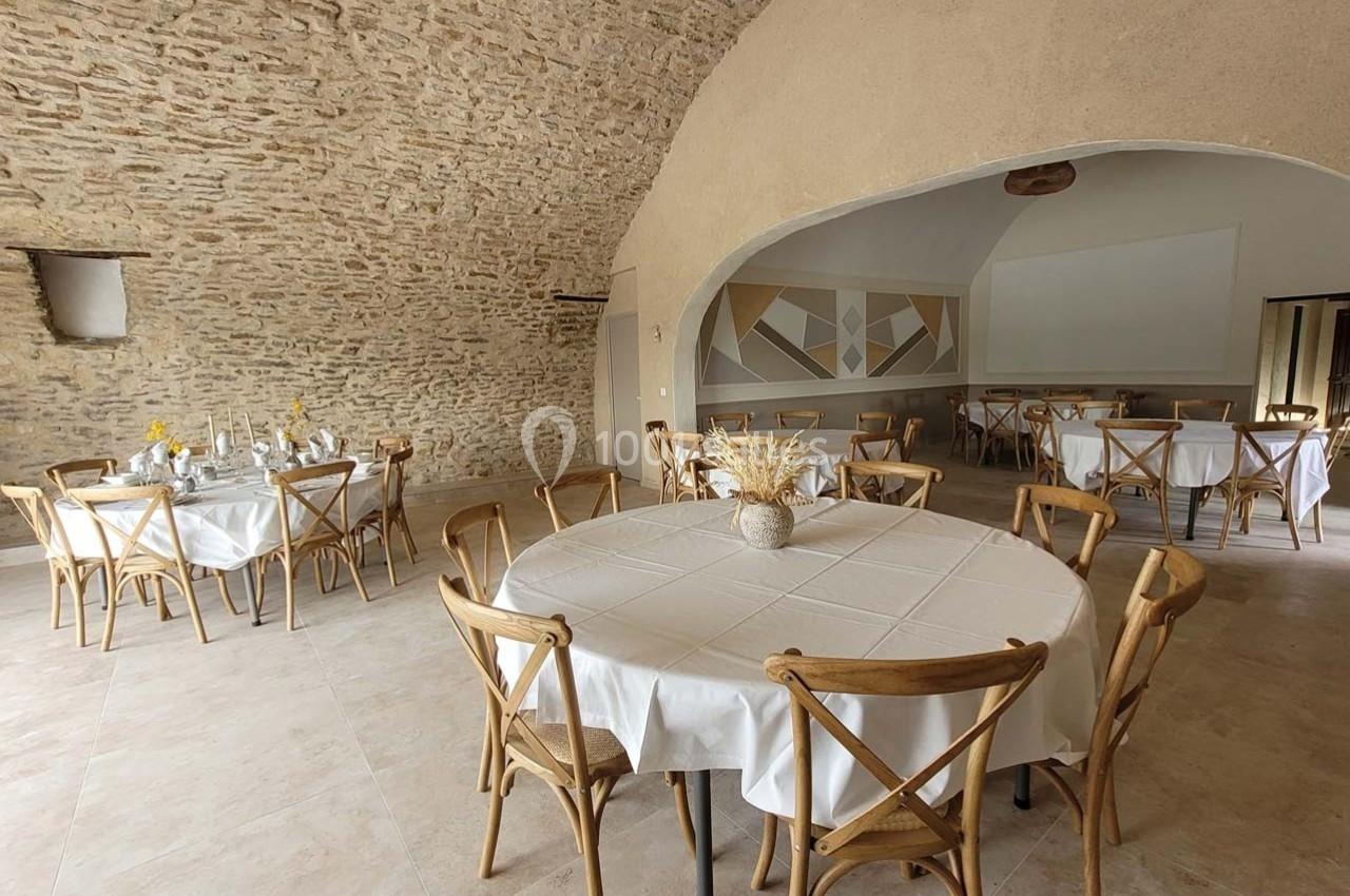 Salle voûtée en pierre avec tables rondes dressées de nappes blanches et chaises en bois, ambiance lumineuse et épurée.