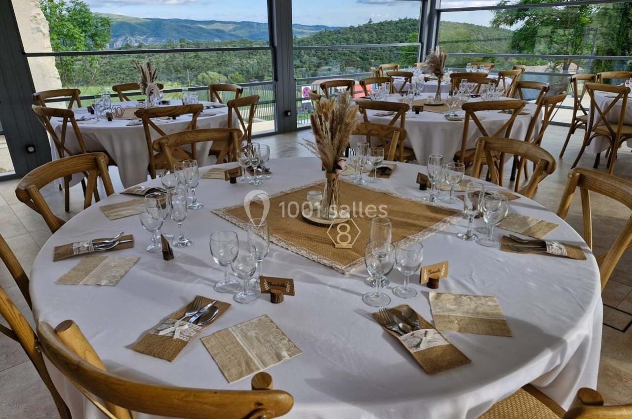 Tables rondes dressées avec nappes blanches, décor champêtre et vue sur un paysage vallonné à travers de grandes baies…