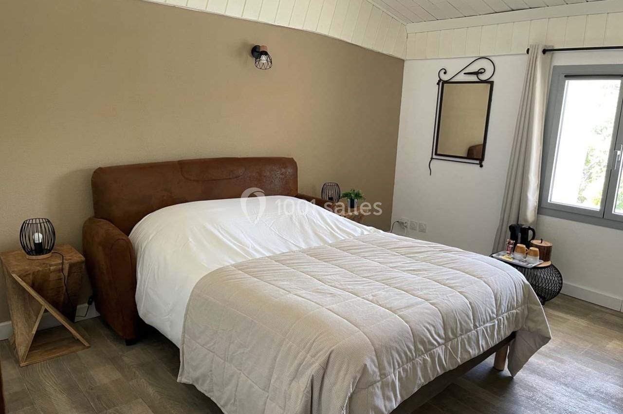 Chambre lumineuse avec lit double, murs beige et blanc, parquet, miroir mural et table de chevet en bois.