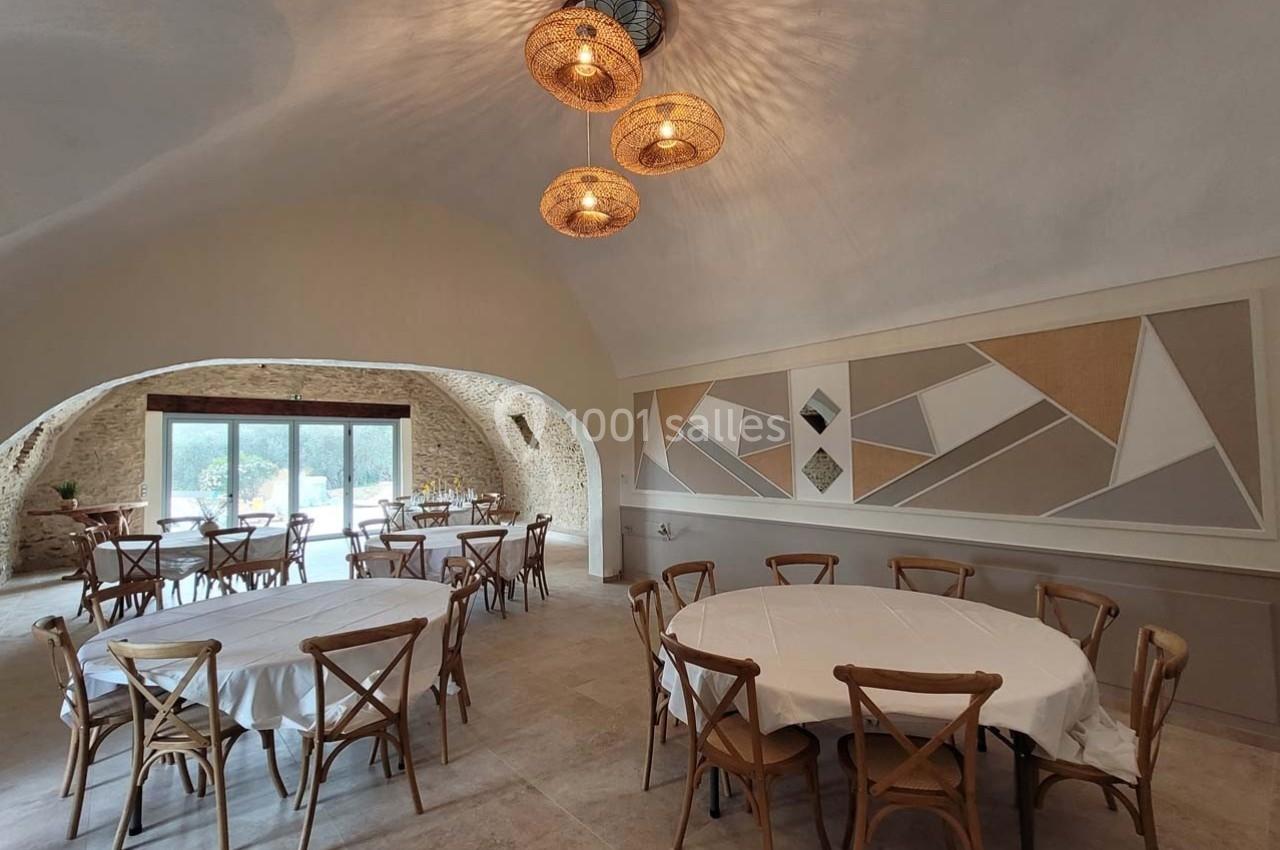 Salle lumineuse avec tables rondes dressées, chaises en bois, murs décorés de motifs géométriques et grandes baies vitrées.