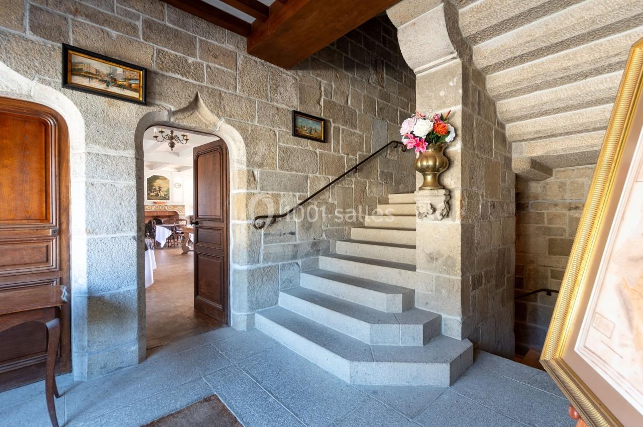 Escalier en pierre dans un intérieur ancien, avec une rampe en métal et un vase de fleurs sur une colonne.