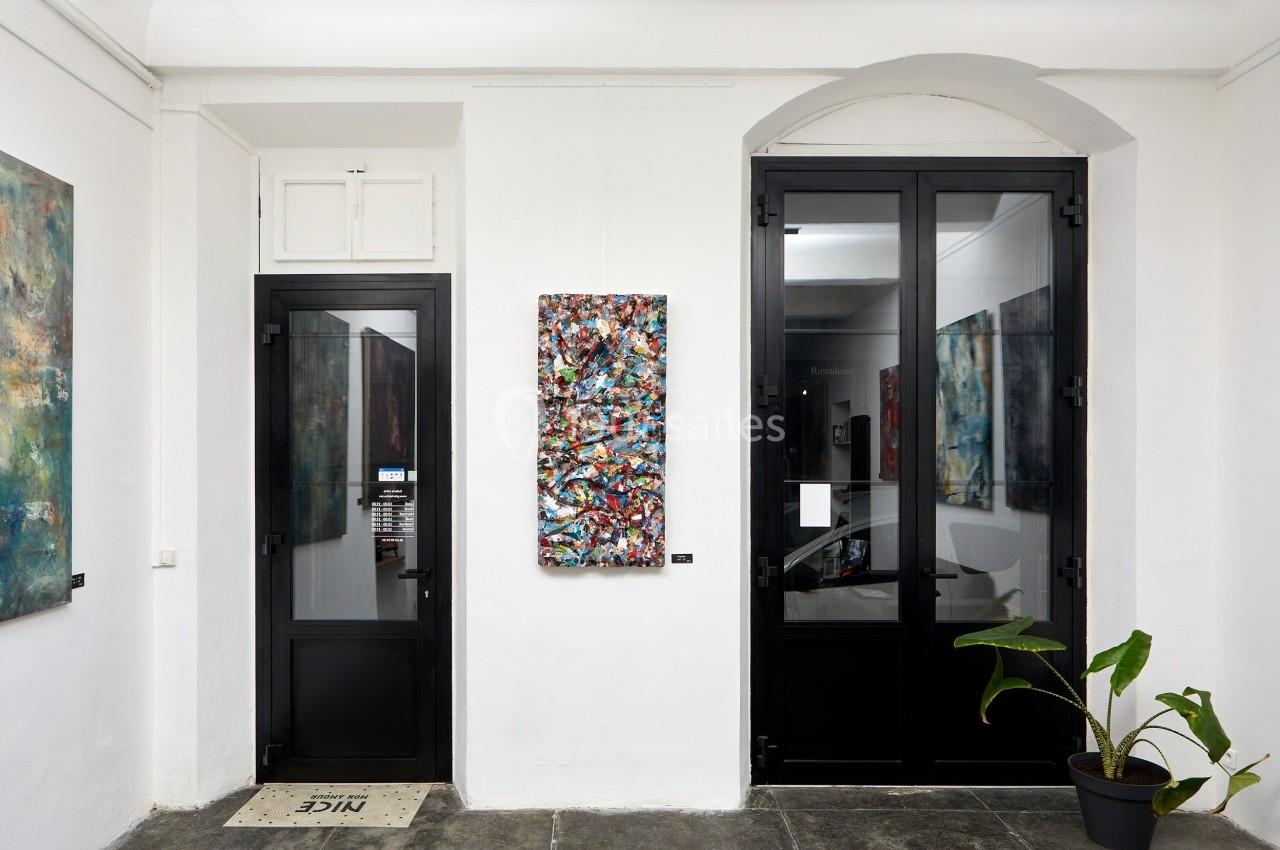 Vue intérieure d'une galerie avec un tableau abstrait coloré accroché sur un mur blanc entre deux portes vitrées.