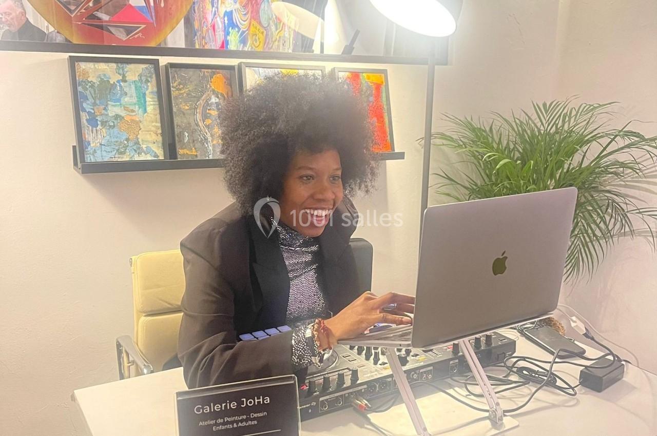 Femme souriante travaillant sur un ordinateur portable dans une galerie d'art, entourée de peintures et d'une plante.