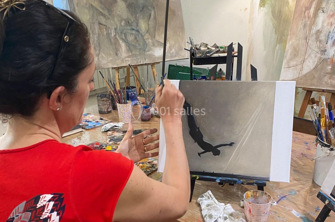 Une femme peint une silhouette sombre sur une toile dans un atelier artistique, entourée de pinceaux et de peintures.