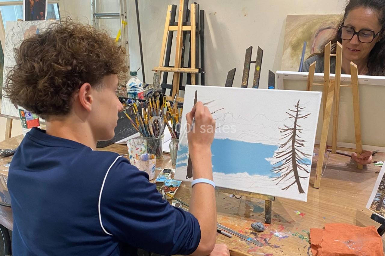 Un jeune homme peint un paysage hivernal sur une toile dans un atelier d'art, accompagné d'une femme en arrière-plan.