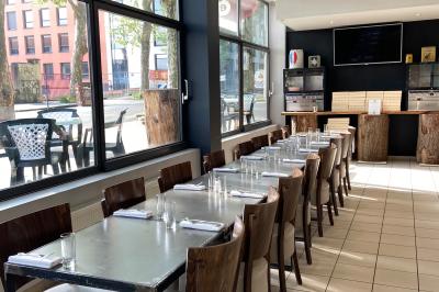 Salle lumineuse avec grandes baies vitrées, tables en bois et vue sur une rue calme bordée de bâtiments.