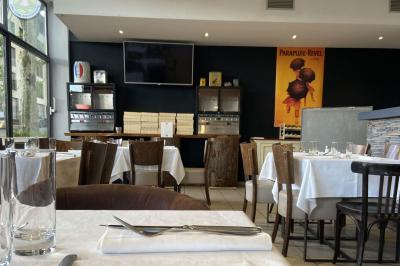 Salle lumineuse avec grandes baies vitrées, tables en bois et vue sur une rue calme bordée de bâtiments.