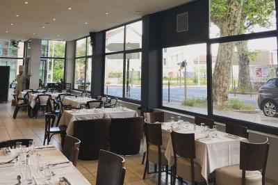 Salle lumineuse avec grandes baies vitrées, tables en bois et vue sur une rue calme bordée de bâtiments.