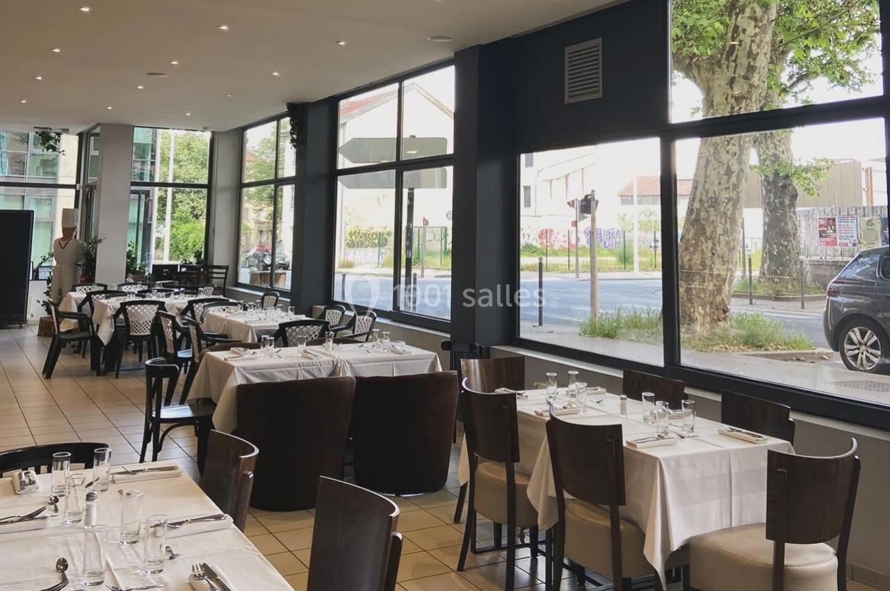 Salle de restaurant lumineuse avec de grandes baies vitrées, des tables dressées et vue sur la rue bordée d'arbres.