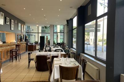 Salle lumineuse avec grandes baies vitrées, tables en bois et vue sur une rue calme bordée de bâtiments.