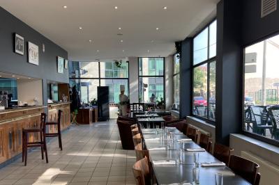 Salle lumineuse avec grandes baies vitrées, tables en bois et vue sur une rue calme bordée de bâtiments.