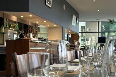 Salle lumineuse avec grandes baies vitrées, tables en bois et vue sur une rue calme bordée de bâtiments.