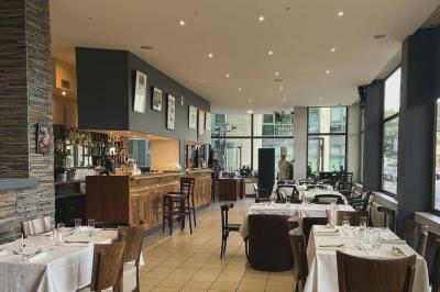 Salle lumineuse avec grandes baies vitrées, tables en bois et vue sur une rue calme bordée de bâtiments.