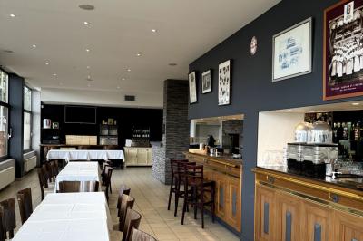 Salle lumineuse avec grandes baies vitrées, tables en bois et vue sur une rue calme bordée de bâtiments.