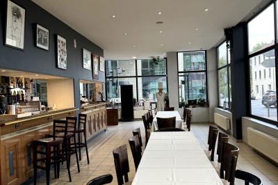 Salle lumineuse avec grandes baies vitrées, tables en bois et vue sur une rue calme bordée de bâtiments.